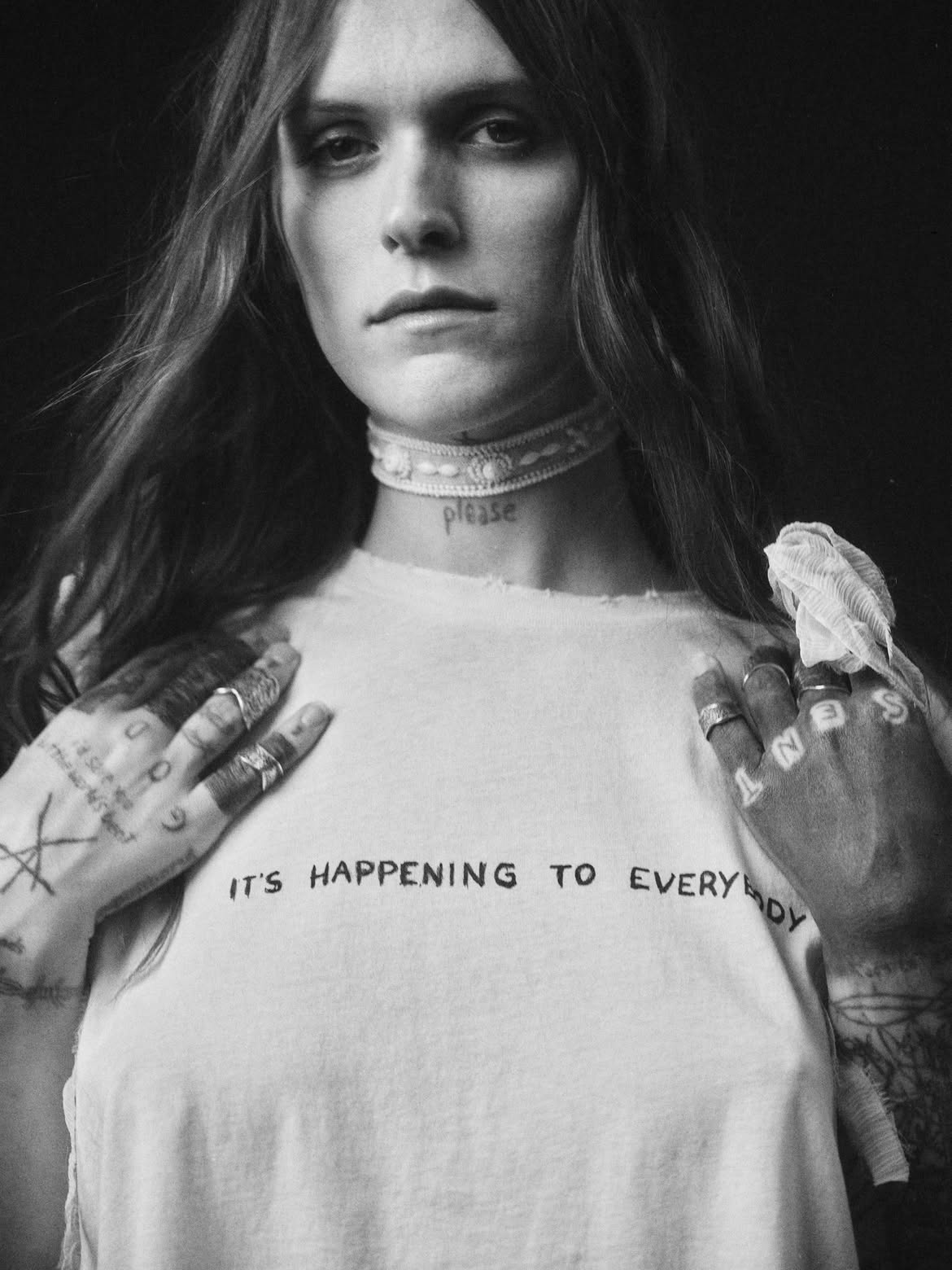 A black and white portrait of a Ethel Cain wearing a light-colored graphic T-shirt with the text 'IT'S HAPPENING TO EVERYBODY' across the chest, set against a dark background for Ann Demeulemeester